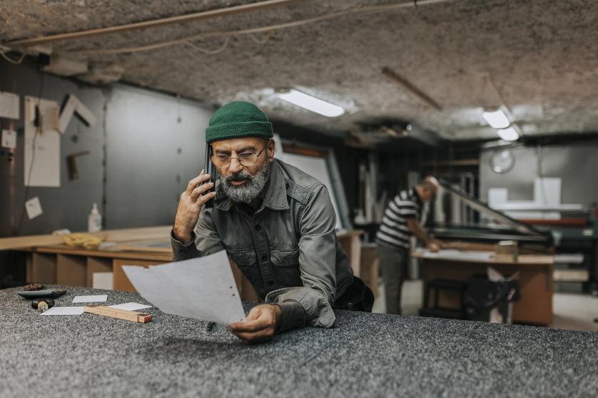 Homem barbudo, proprietário de uma planta, falando ao telefone inteligente enquanto se apoia em uma bancada de trabalho